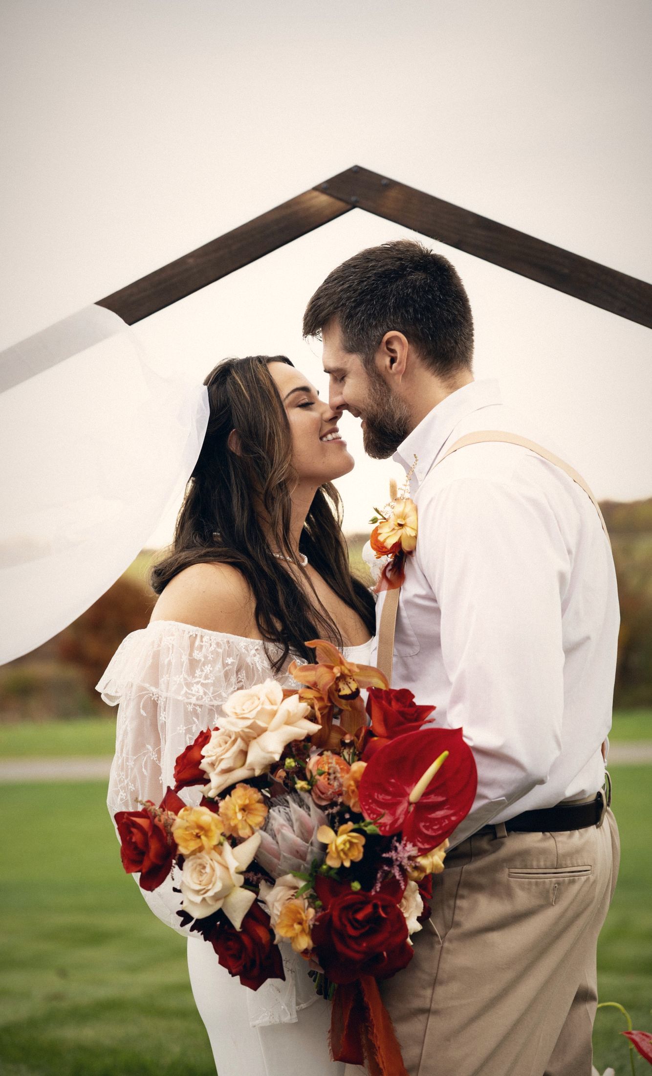 Couple touching noses under a wooden arch with a red bouquet