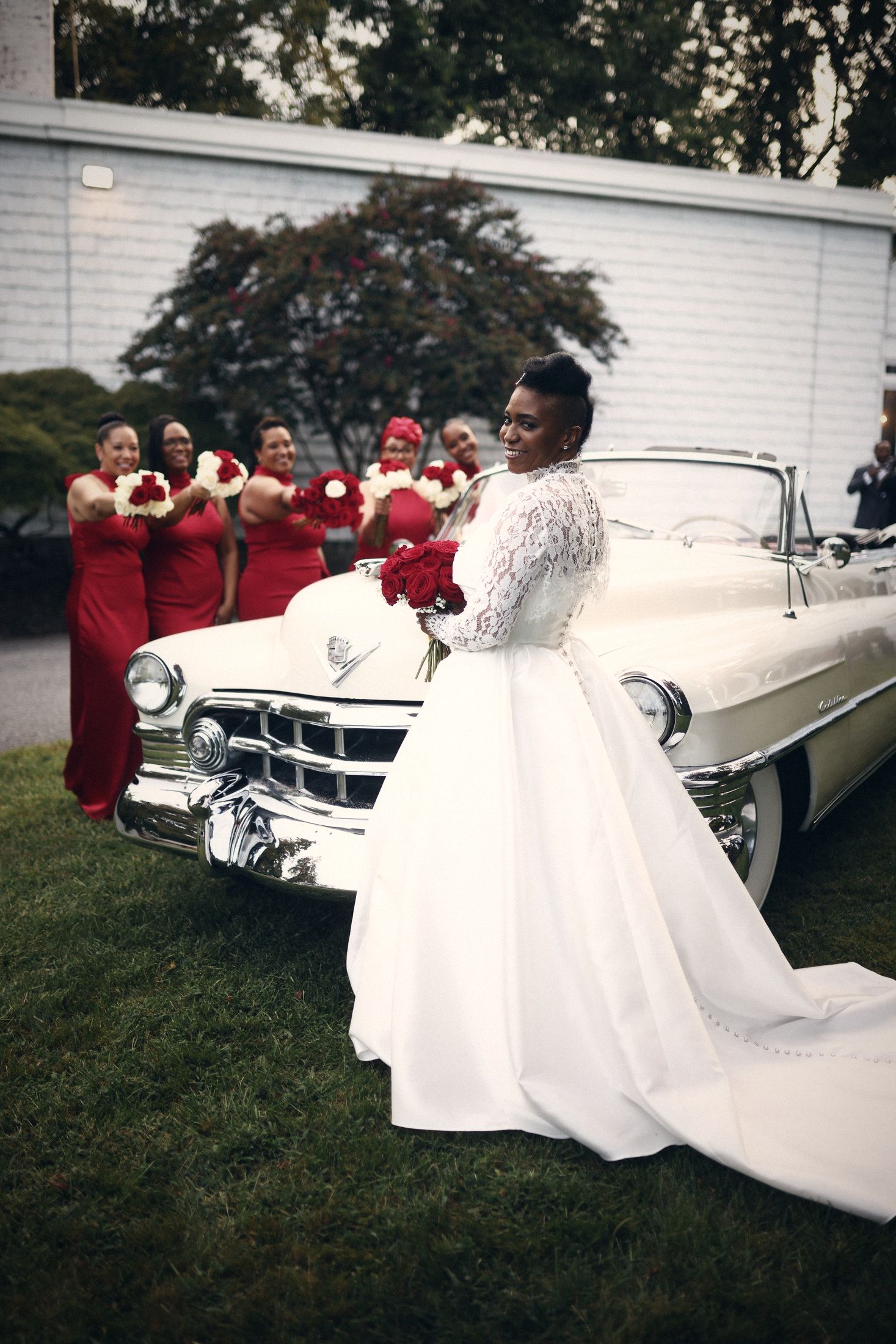 Bride beside a vintage convertible with bridesmaids in red