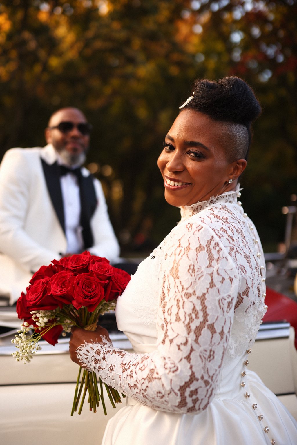Bride glancing back with red roses as the groom waits in a vintage convertible
