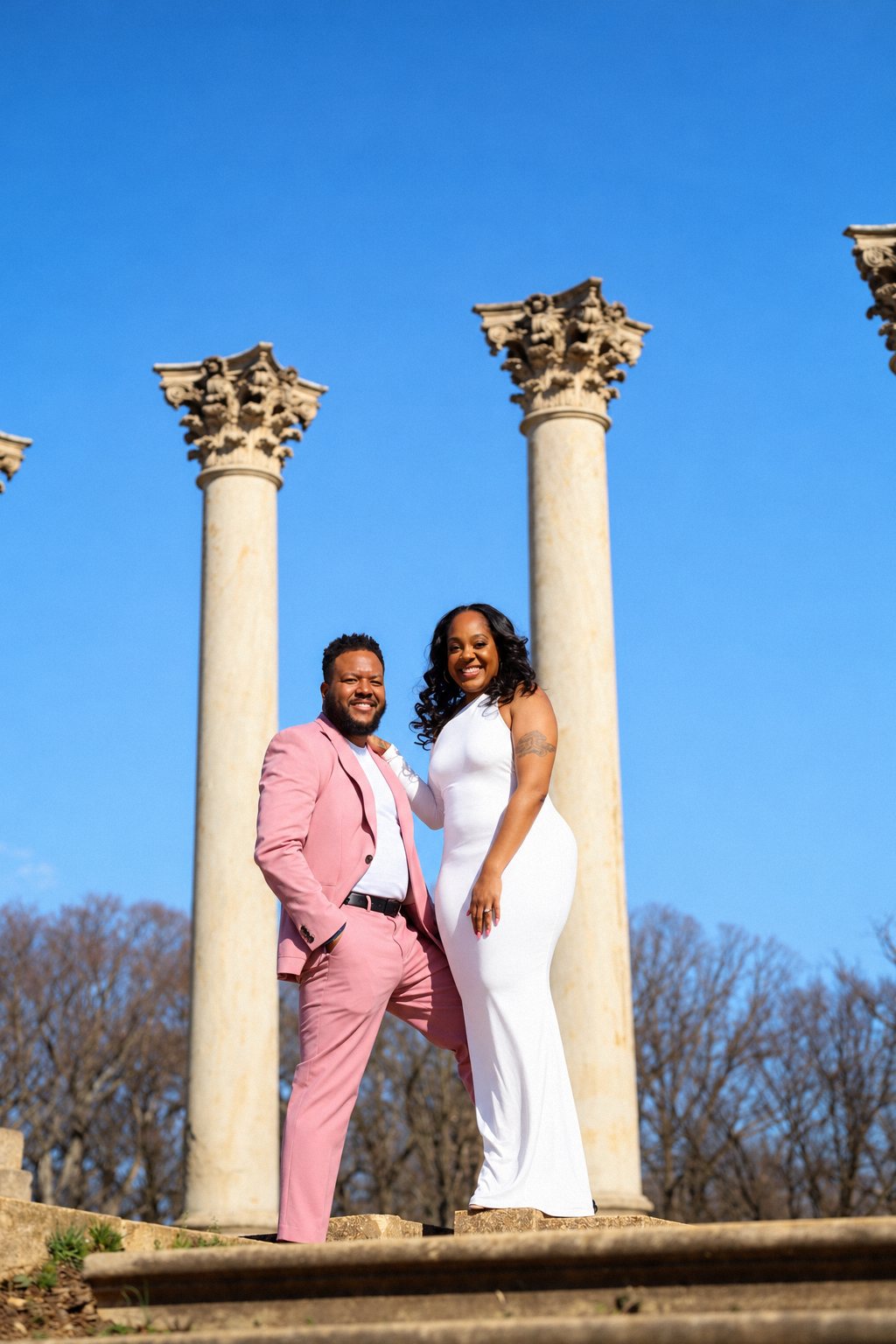 Couple in a pink suit and white dress posing between marble columns