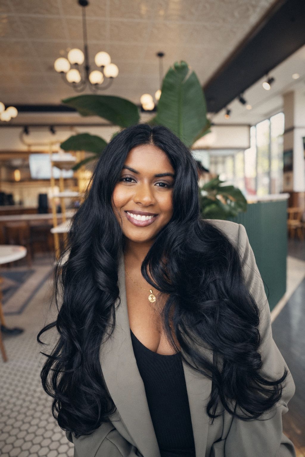 Smiling woman with long dark hair in an olive blazer at a cafe