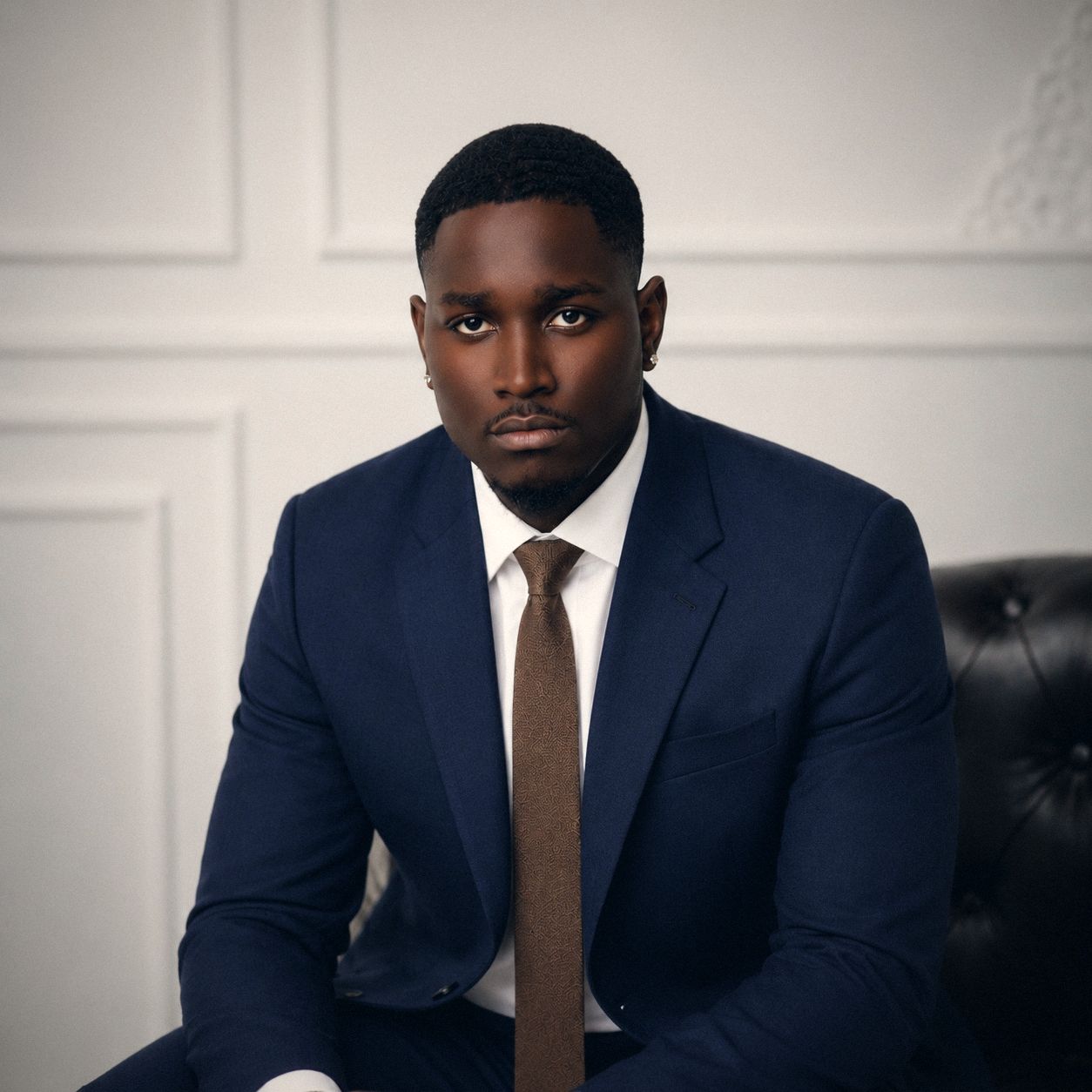 Man in a navy suit and brown tie seated against a white paneled wall