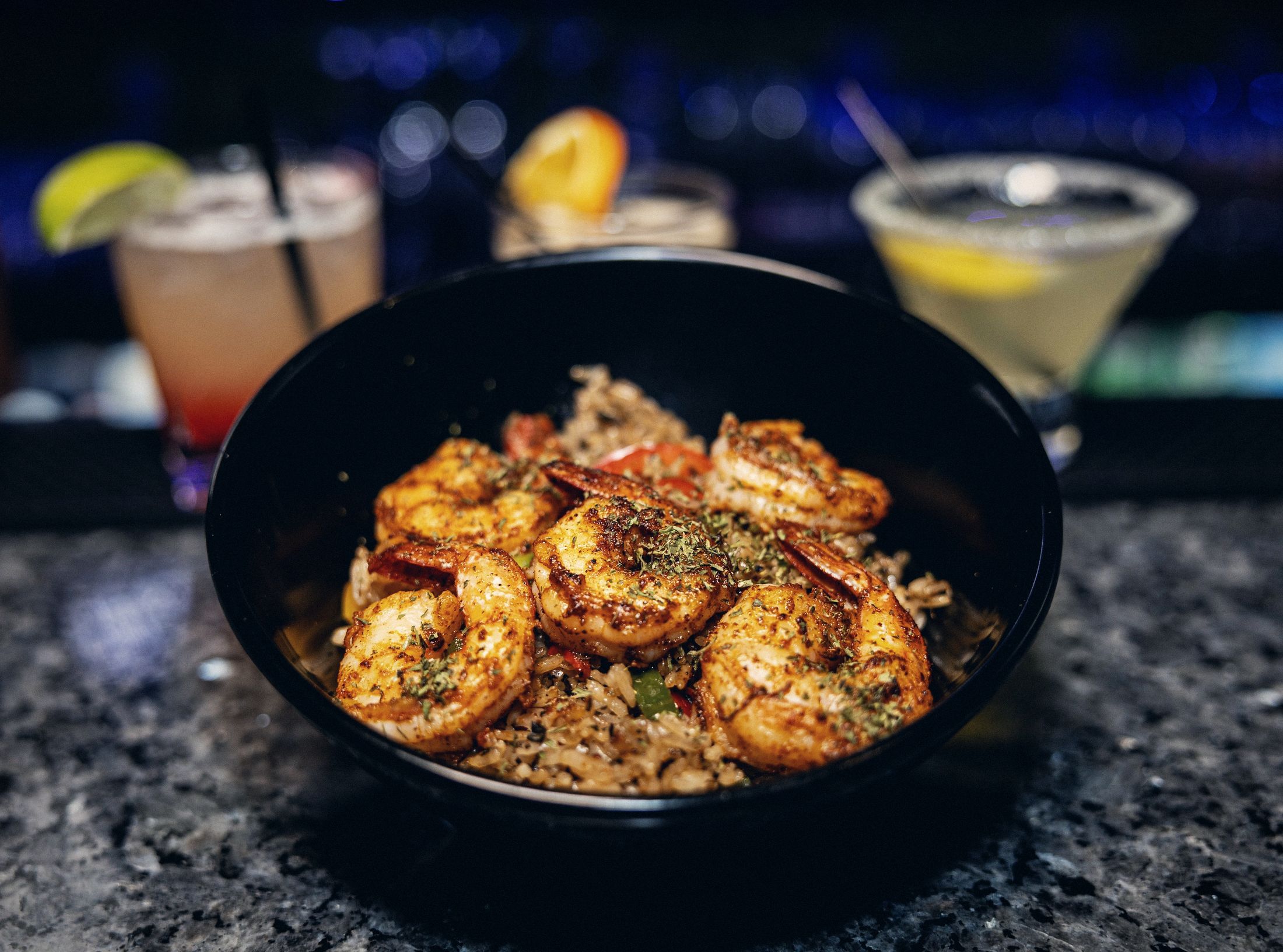 Seasoned shrimp over rice in a black bowl with cocktails behind