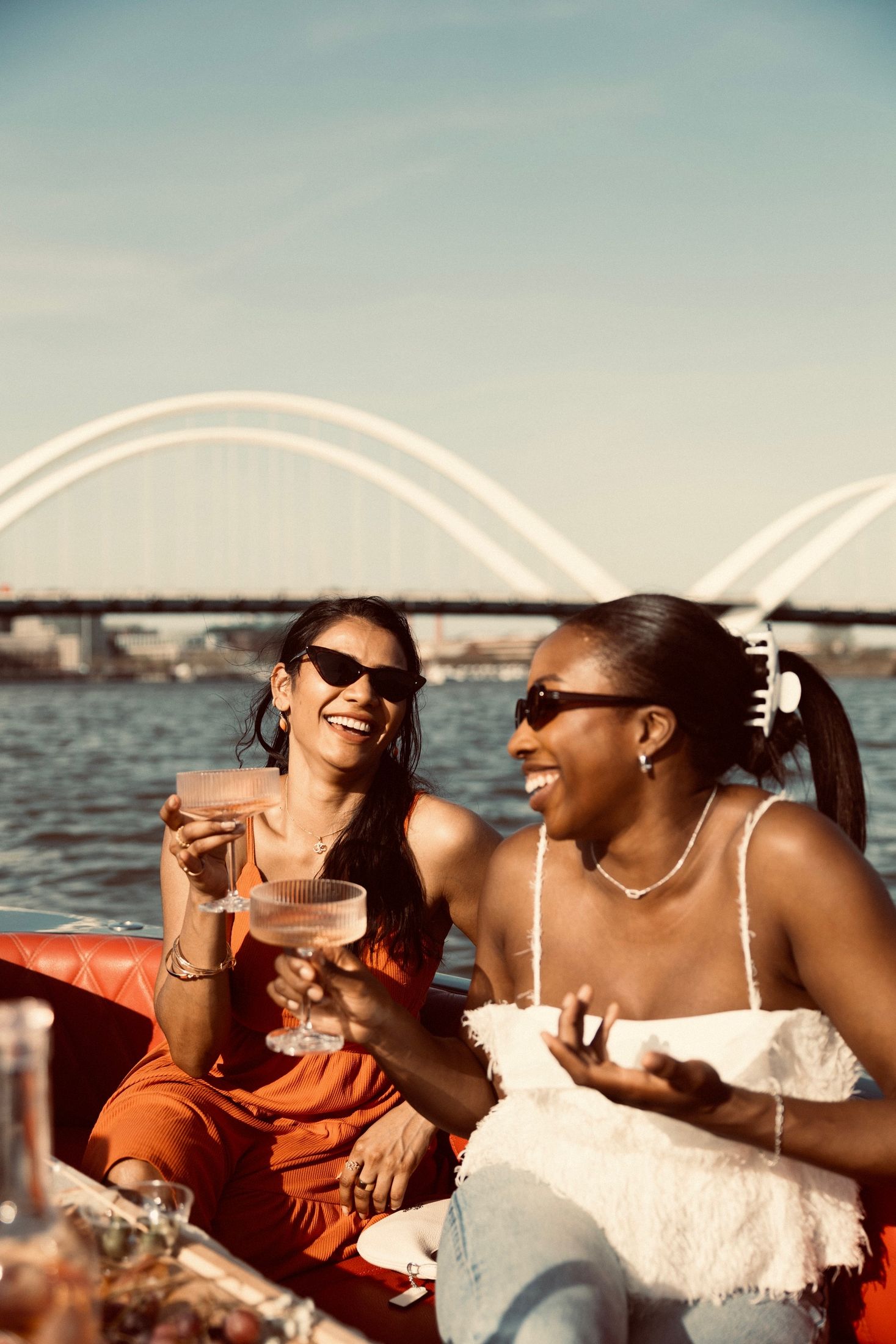 Two friends laughing with drinks on a boat by the Frederick Douglass Bridge